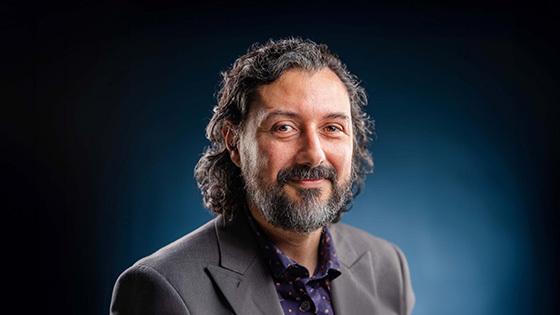 Plinio Pinto smiles at the camera. He has curly brown hair and a beard streaked with gray, and he wears a gray blazer.